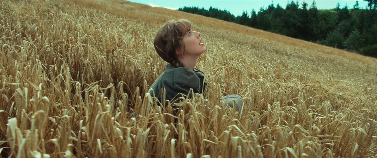 Sunset Song (2015) movie still: medium — A woman kneels amidst a dense field of tall, dry, golden-brown wheat stalks. She stares upward toward the…