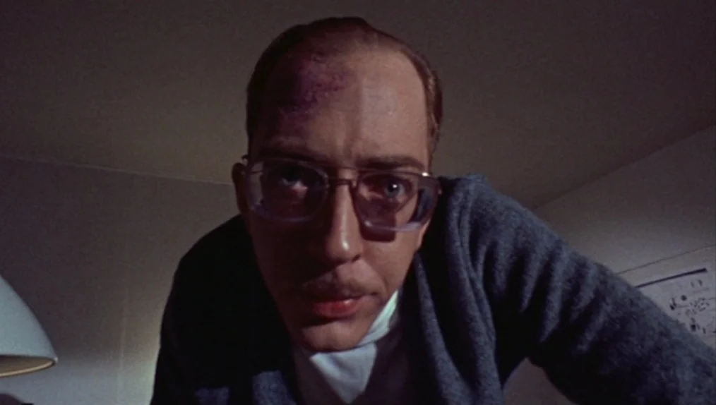 Sisters (1973) movie still: closeup — A dimly lit room with plain walls frames a man from a low angle. The man leans over the lens while looking…