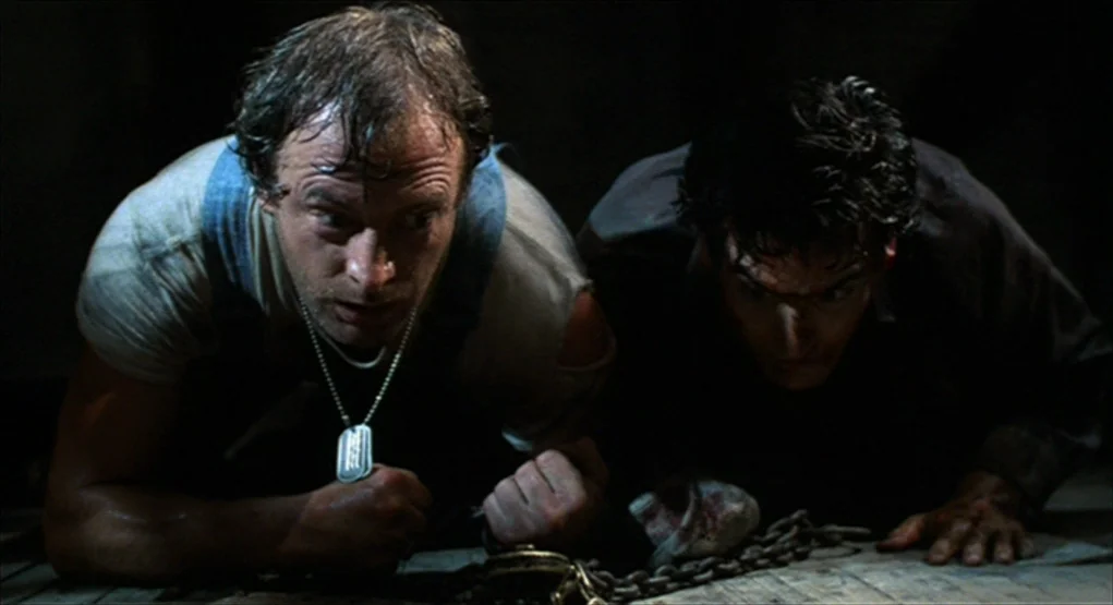 Evil Dead II (1987) movie still: medium — A dimly lit, claustrophobic environment with a weathered wooden floor. Two men crawl forward while…
