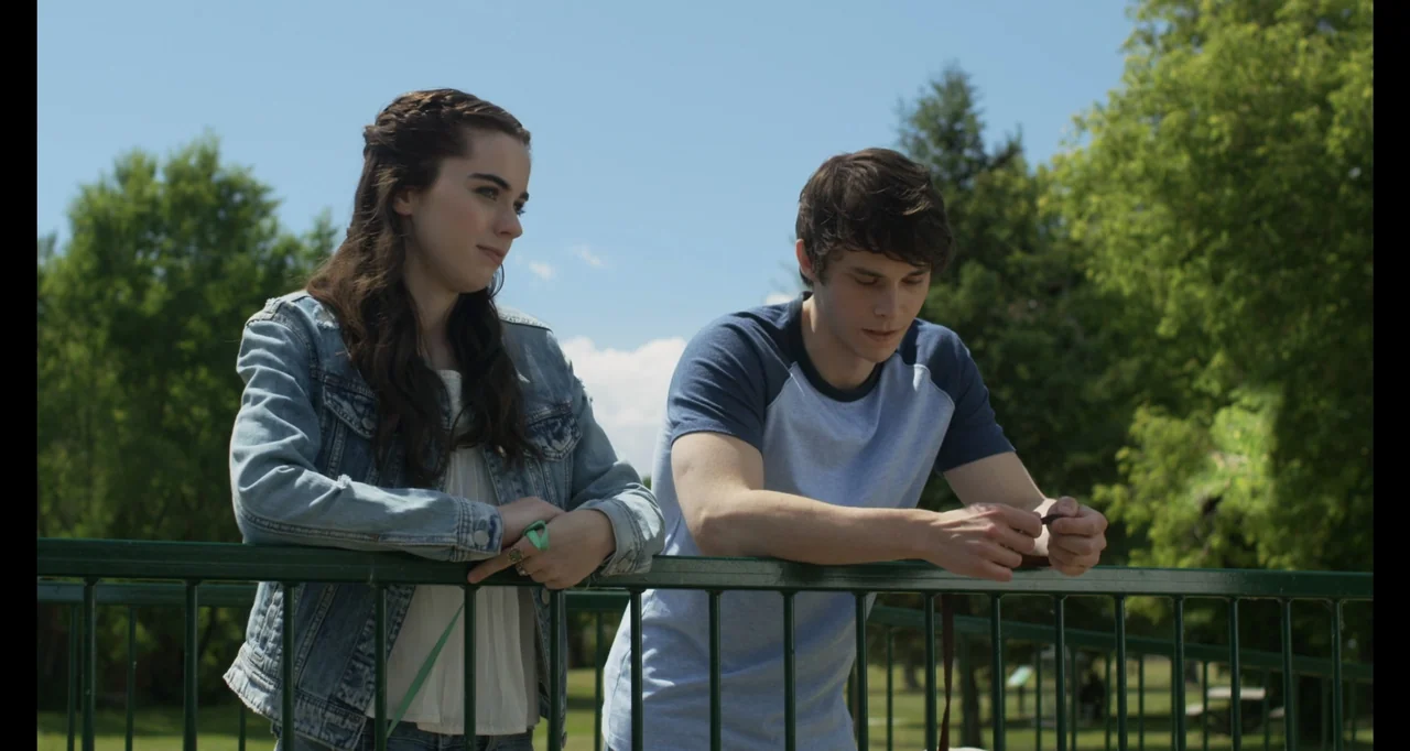 A.R.C.H.I.E. (2016) movie still: medium — Two young adults stand side by side on a green metal bridge in a park. They lean against the railing while…