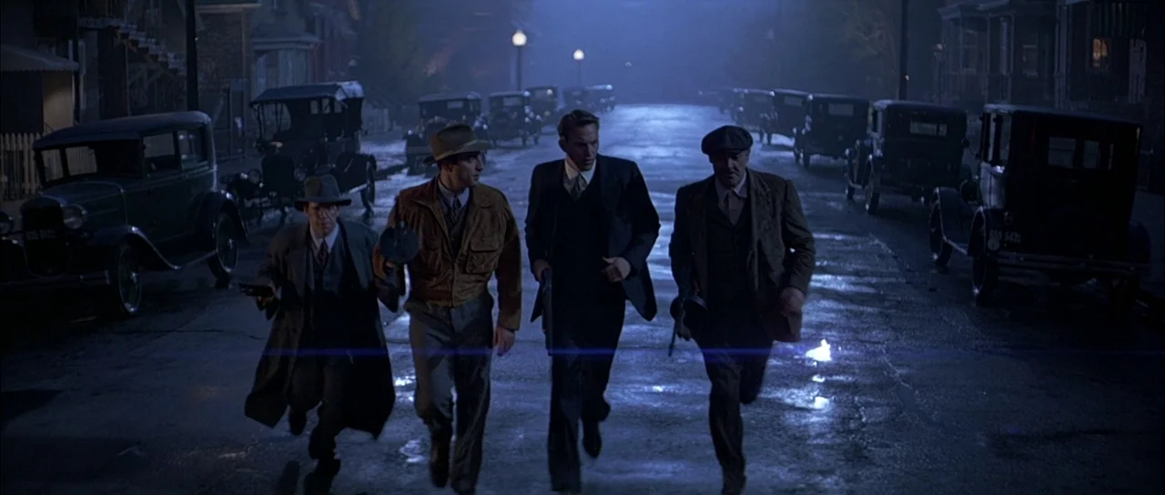 The Untouchables (1987) movie still: fullbody — A wet 1930s cobblestone street is lined with multiple vintage parked automobiles under lamps. Four…
