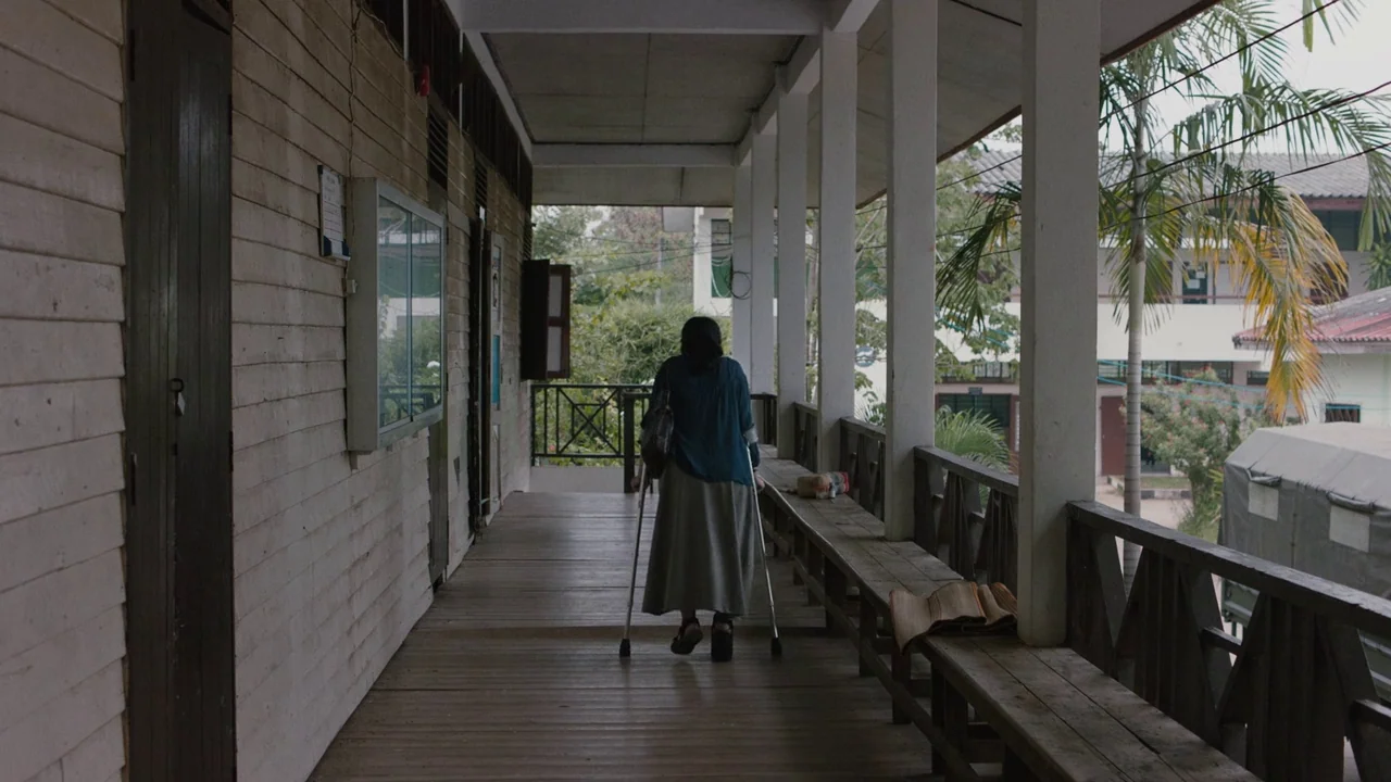 Cemetery of Splendour (2015) movie still: fullbody — A wooden porch at a school features white pillars and a long bench. A person in a blue shirt…