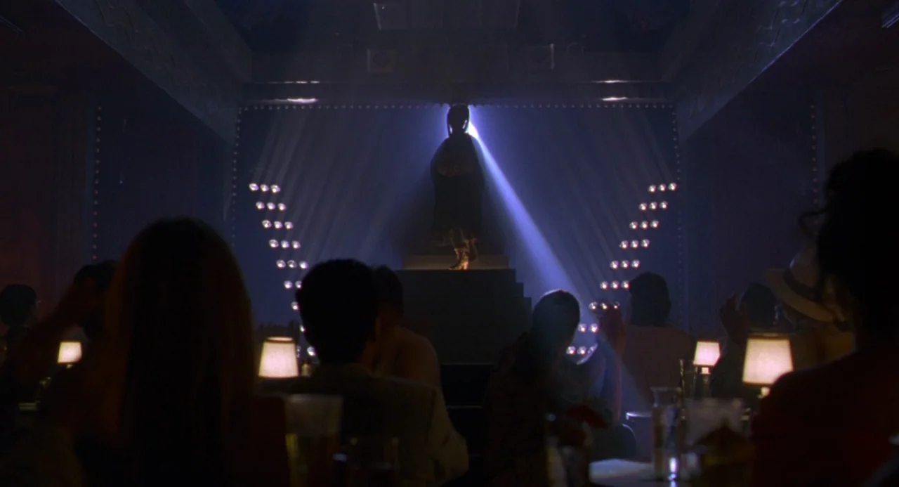 The Birdcage (1996) movie still: wide — A dimly lit club with audience members seated at tables facing a central stage. A lone performer stands as a…