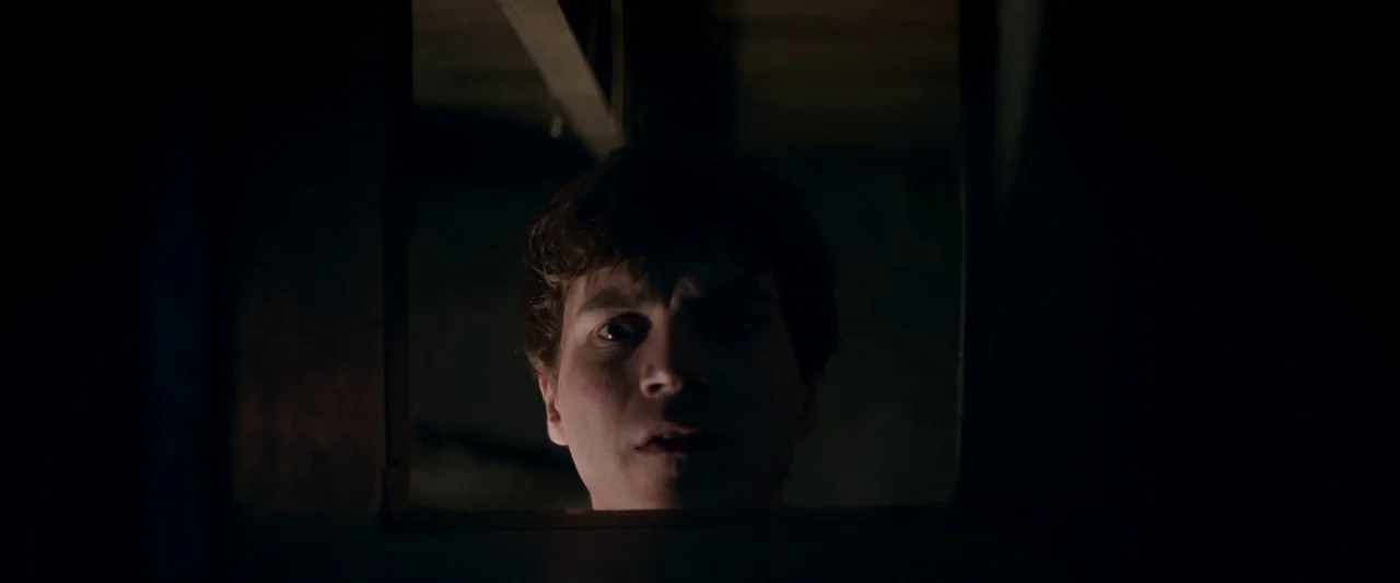 The Autopsy of Jane Doe (2016) movie still: closeup — A dark, cramped space frames the head and shoulders of a young man. He looks through a…