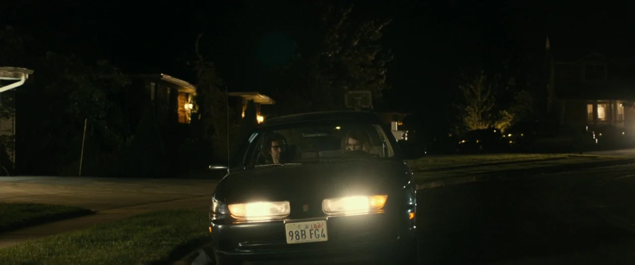 Brigsby Bear (2017) movie still: wide — A dark suburban residential street at night features a sedan parked at the curb. Two people sit inside the…