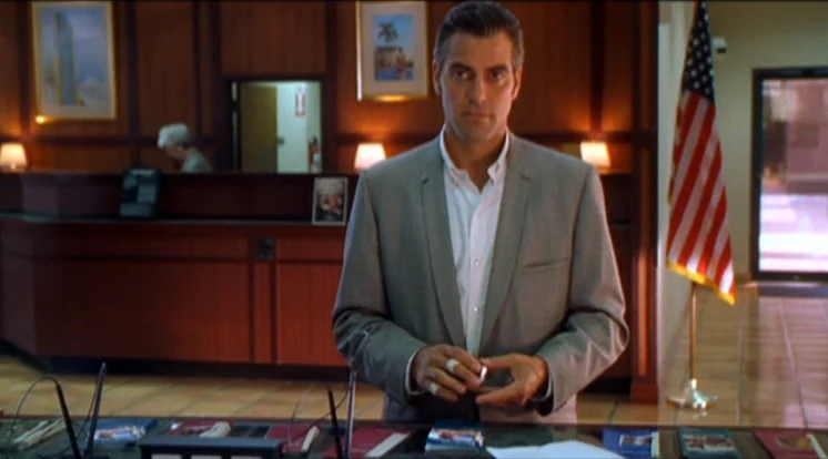 Out Of Sight (1998) movie still: medium — A man stands in front of a wood-paneled wall decorated with framed pictures. The man performs a coin…
