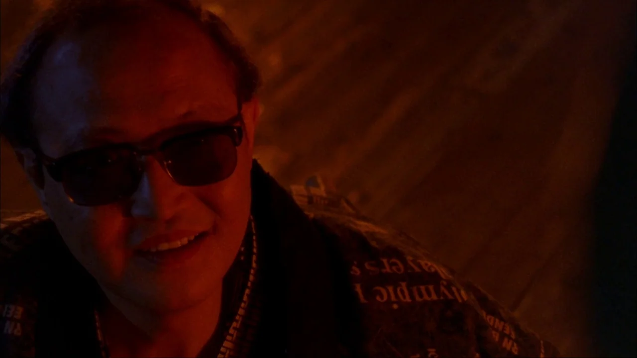 Audition (1999) movie still: closeup — An interior space lit by dim red lighting. A man wearing glasses smiles while looking toward the side of the…