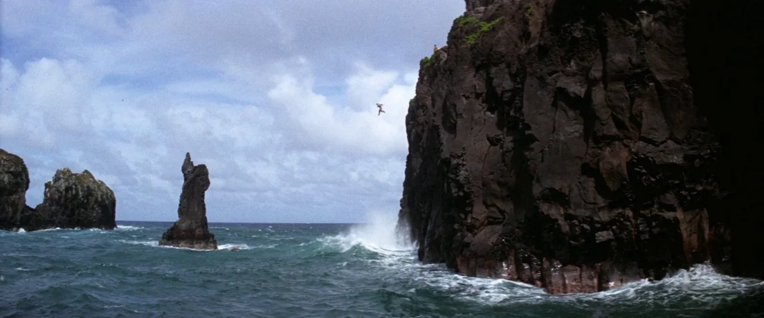 Papillon (1973) movie still: wide — Jagged volcanic rock cliffs rise vertically from churning ocean waves under a cloudy sky. A small figure jumps…
