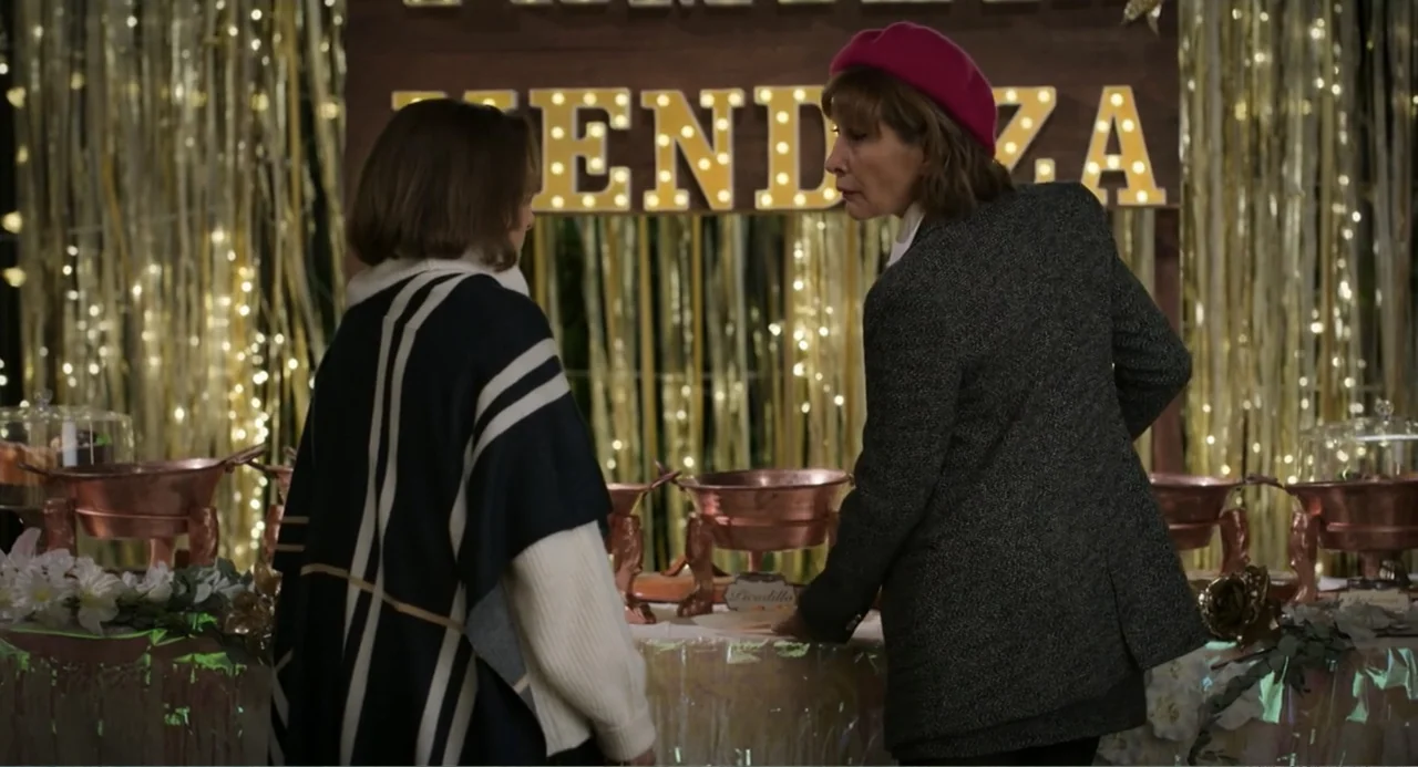 La posada (2023) movie still: medium — A festive buffet display features copper warming dishes and string lights mounted on a wall. Two women stand…