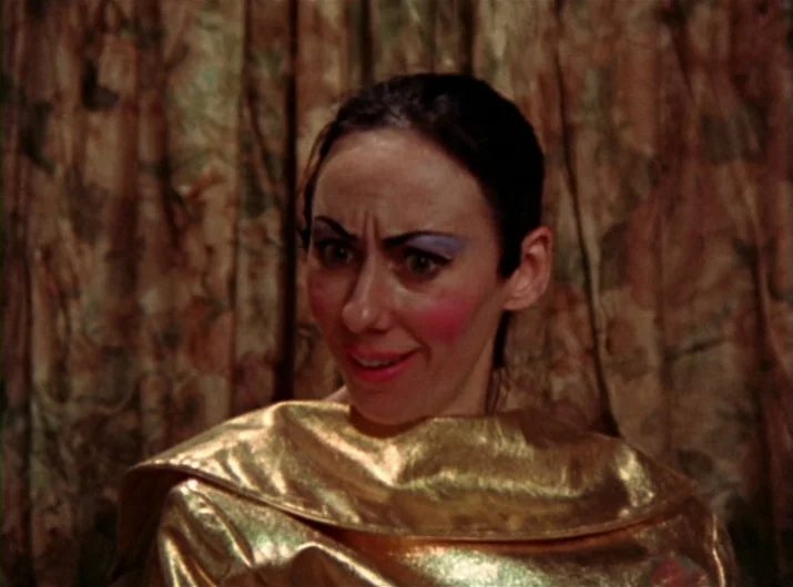 Desperate Living (1977) movie still: closeup — A woman stands in front of a patterned fabric backdrop in a room. She wears a shiny gold top and…