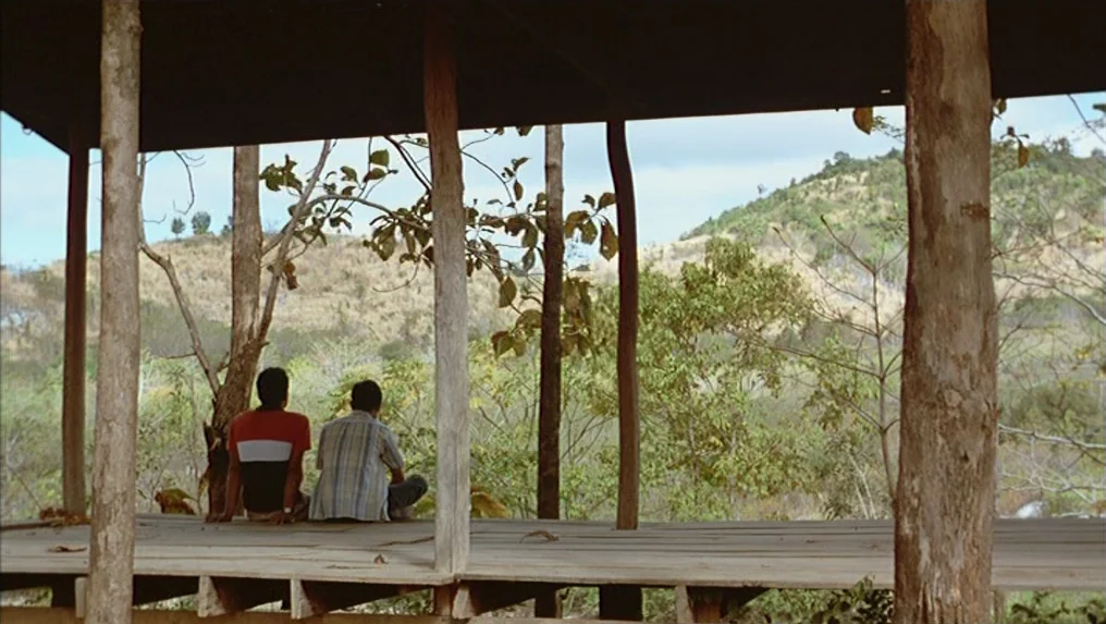 Tropical Malady (2004) movie still: wide — A wooden platform with vertical support posts frames an open view of hills. Two people are seated on the…