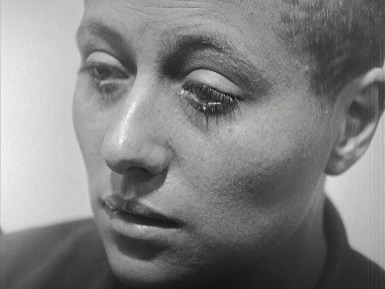Vivre Sa Vie (1962) movie still: closeup — A close-up of a woman with shaved hair against a light wall. She stares downward with eyelids partially…