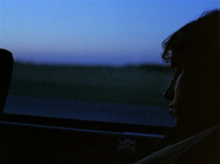 Slacker (1991) movie still: closeup — The interior of a car at twilight with a dark, blurred exterior. A young man sits in profile looking out the…