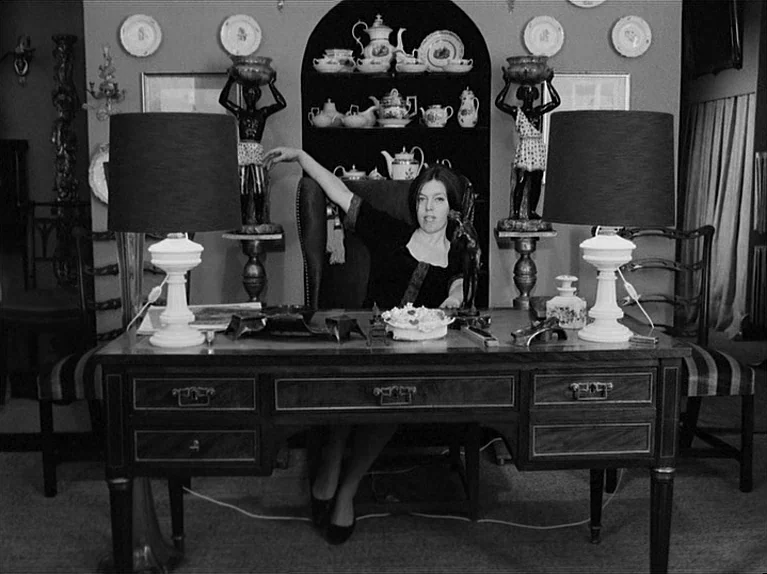 Gods Of The Plague (1970) movie still: medium — A woman sits at an ornate wooden desk centered between two decorative black statues. Plates hang on…