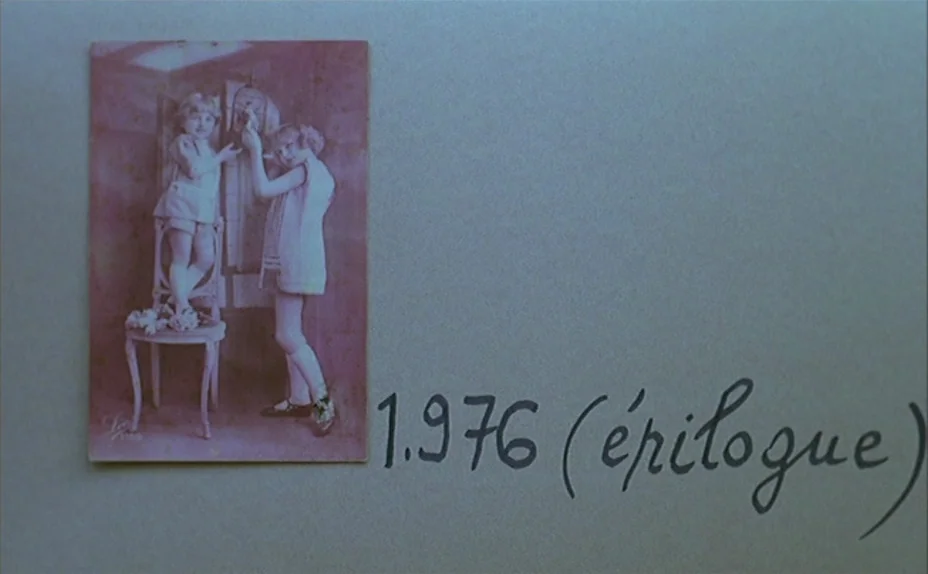 L’une Chante, l’Autre Pas (1977) movie still: medium — A vintage sepia-toned photograph of two children is pinned to a plain gray surface.…