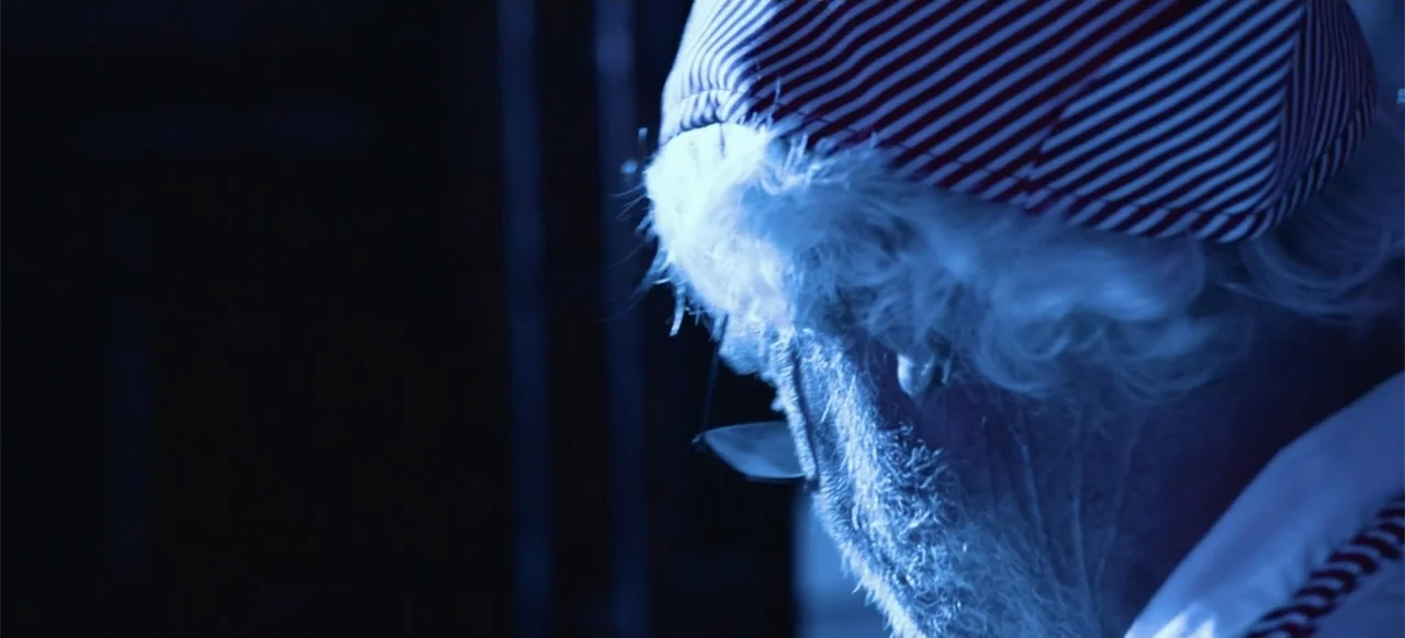 Hellbreeder (2004) movie still: closeup — A dark indoor space contains an elderly man with a white beard. The man wears a red and white striped cap…