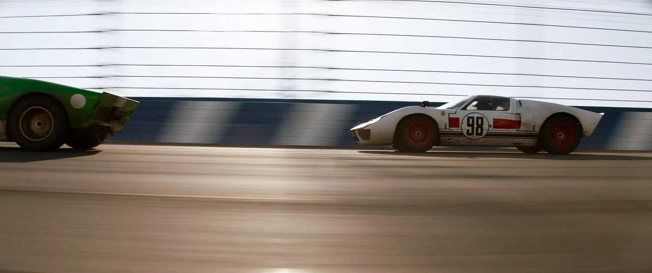 Ford V Ferrari (2019) movie still: wide — A banked asphalt racetrack features a tall safety fence against a bright sky. A white Ford GT40 with the…