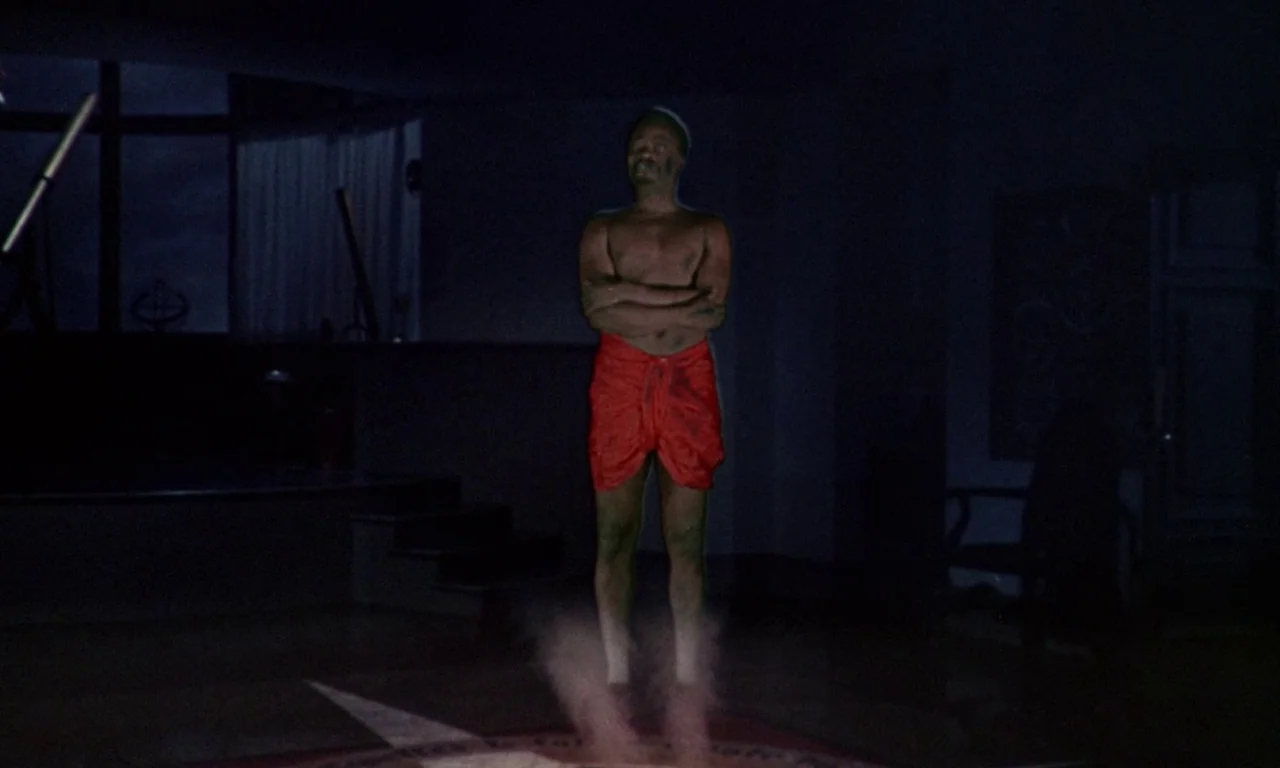 The Devil Rides Out (1968) movie still: fullbody — A darkened, sparse room contains a faint dust cloud at the floor level. A man floats inches above…