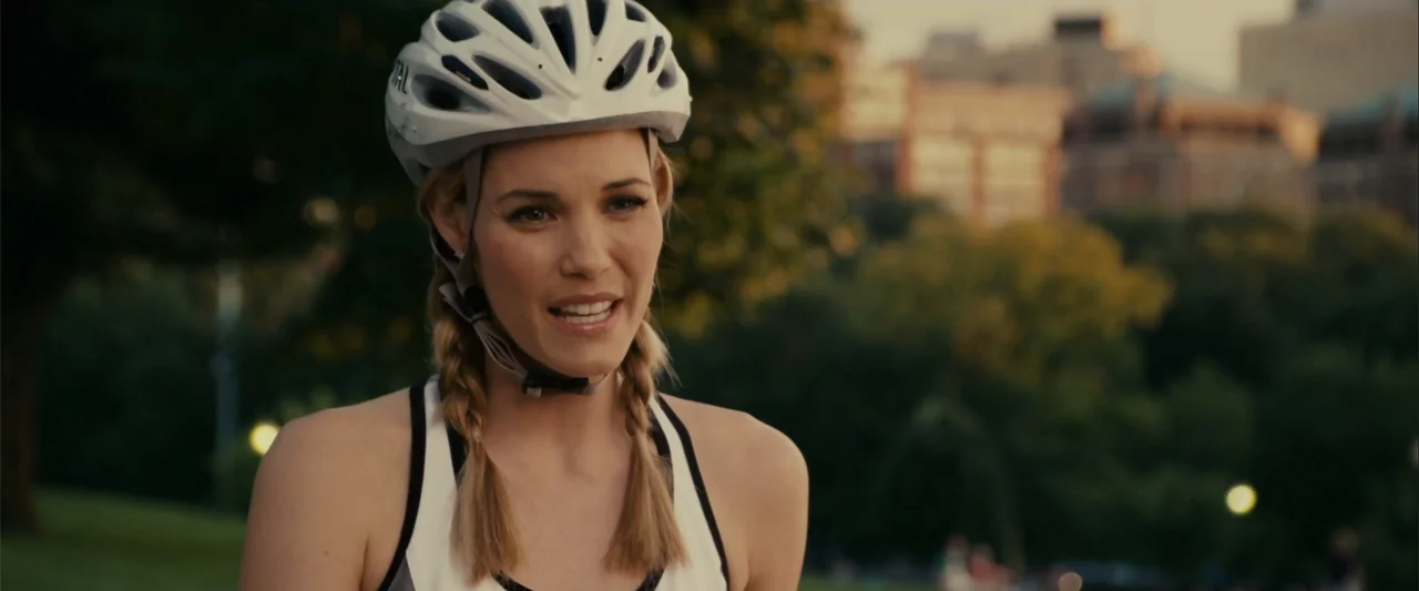 Zookeeper (2011) movie still: medium — A park setting with blurred greenery and urban architecture in the distant background. A woman in a white…