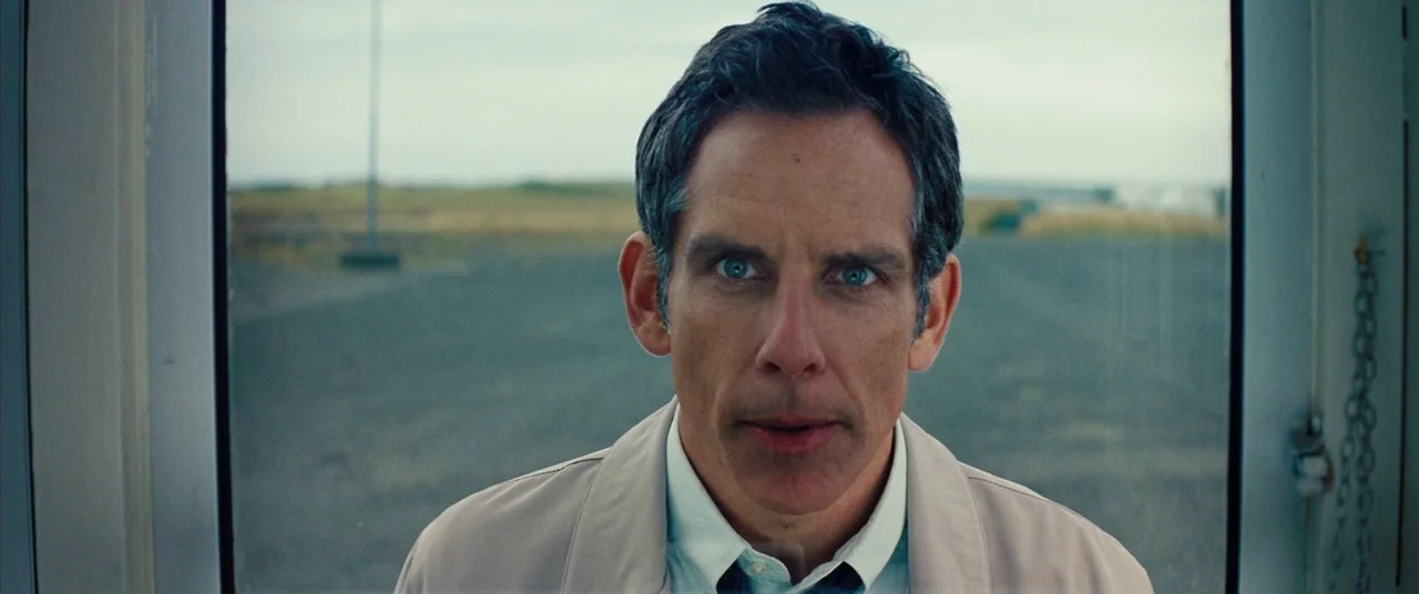 The Secret Life of Walter Mitty (2013) movie still: closeup — A man looks through a glass window at an overcast outdoor field. He stands framed…
