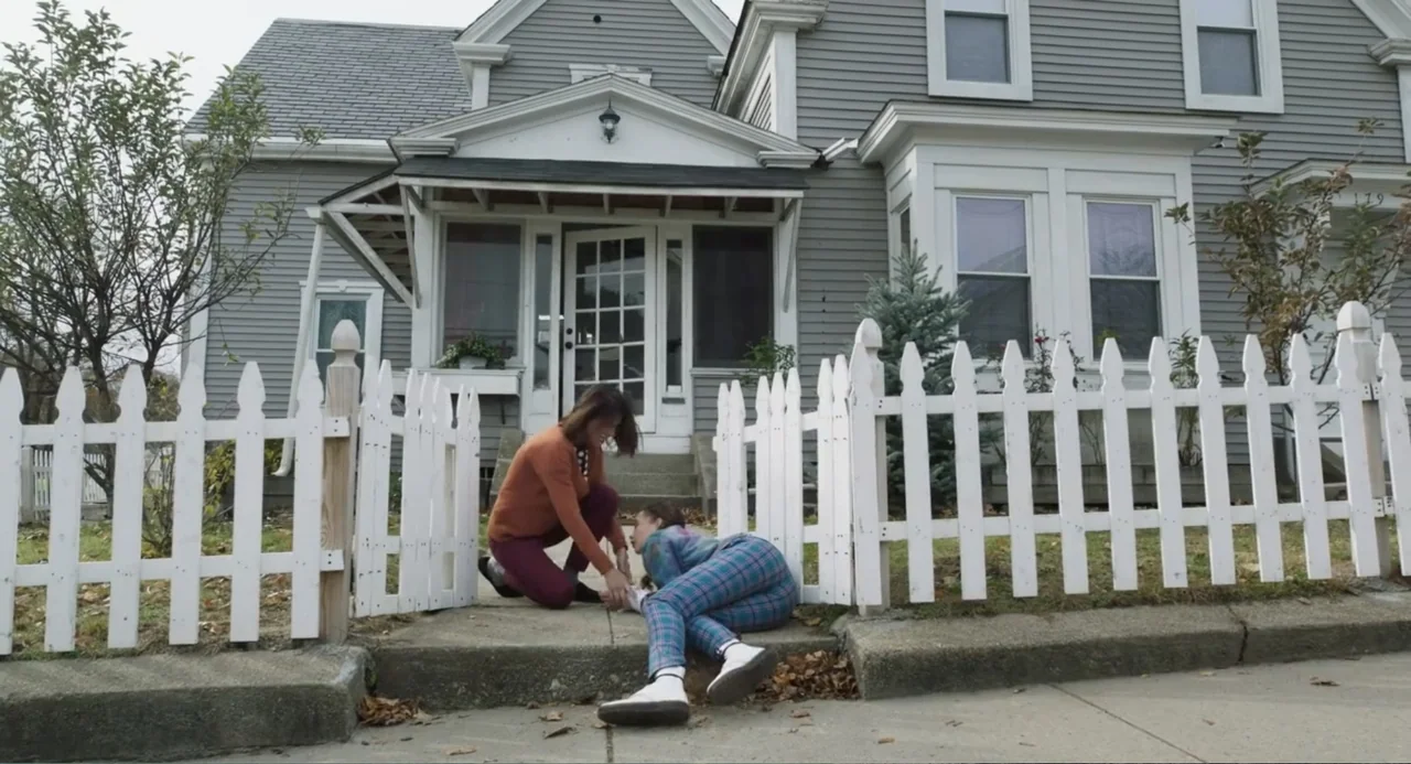 Salvation (2025) movie still: medium — A suburban gray house stands behind a white picket fence with a paved walkway. A woman kneels on the ground…