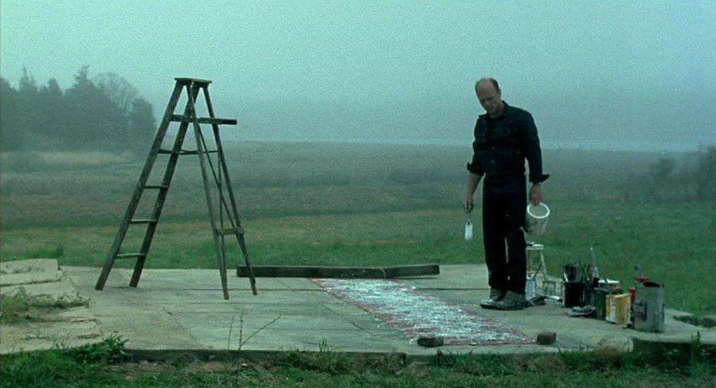 Pollock (2000) movie still: wide — An open grassy field features a concrete platform set against a dense fog background. A middle-aged man stands…
