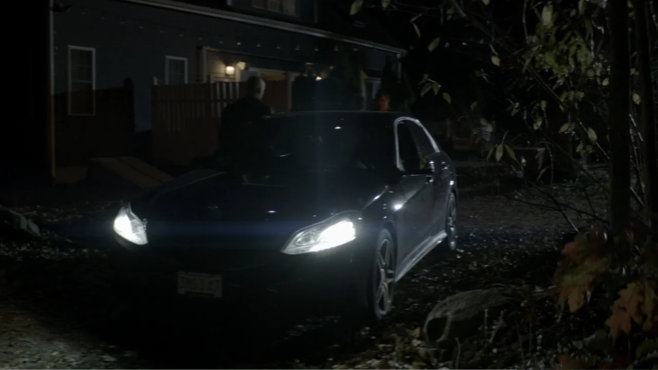 The Collective (2023) movie still: medium — A dark outdoor driveway at night features a sedan parked with headlights shining bright. A person stands…