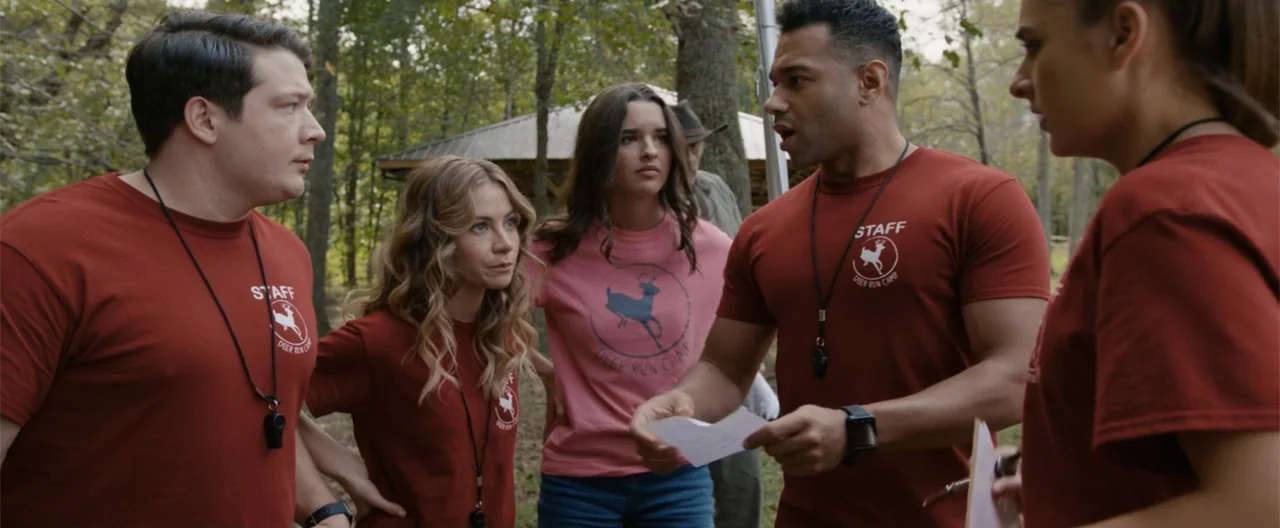 Camp Hideout (2023) movie still: medium — A wooded outdoor camp area with mature trees and a gazebo structure visible behind. Four staff members in…