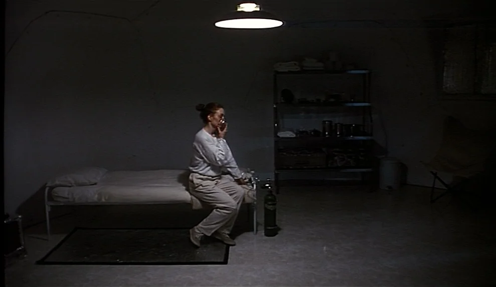 Safe movie still: wide — A dark subterranean bunker with bare concrete walls and minimal institutional furnishings. A woman sits on a thin cot while…