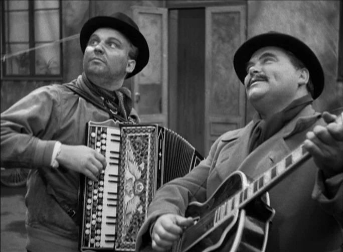 Summer with Monika (1953) movie still: medium — An outdoor courtyard with rustic plaster walls and a paneled doorway in the background. Two men in…