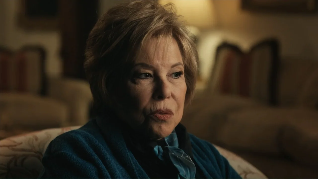 The Winding Stream (2014) movie still: closeup — A dimly lit room with soft furniture and muted tones. An older woman with blonde hair sits while…