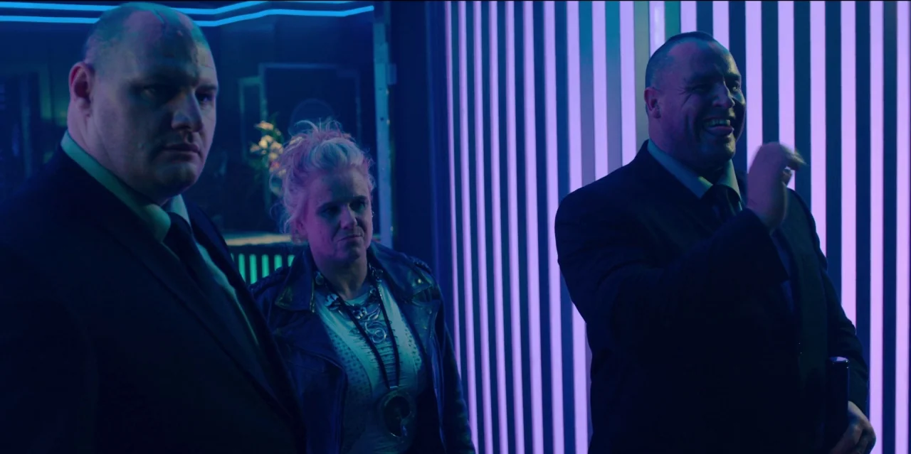 Mute (2018) movie still: medium — A dark venue interior features walls lined with bright, vertical purple neon lighting strips. Three people stand…