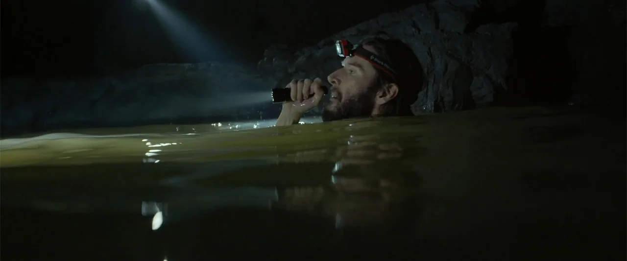 Black Water: Abyss (2020) movie still: medium — A man is chest-deep in dark murky water inside a damp rocky cave. He holds a bright flashlight and…