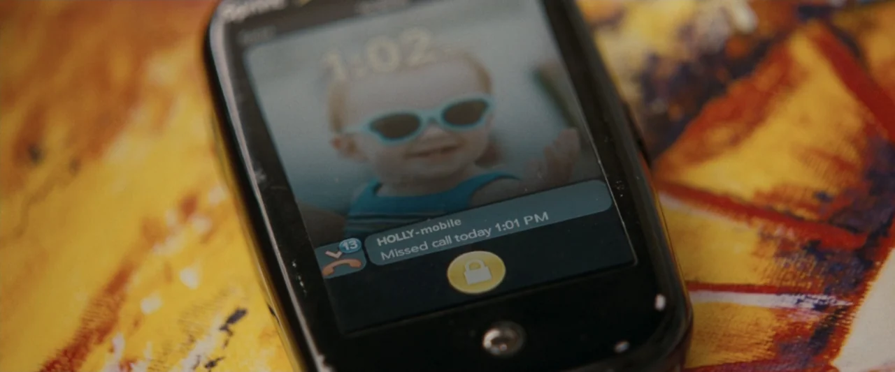 Life As We Know It (2010) movie still: closeup — A close-up shows a black smartphone resting on a vibrant, multi-colored patterned fabric surface.…