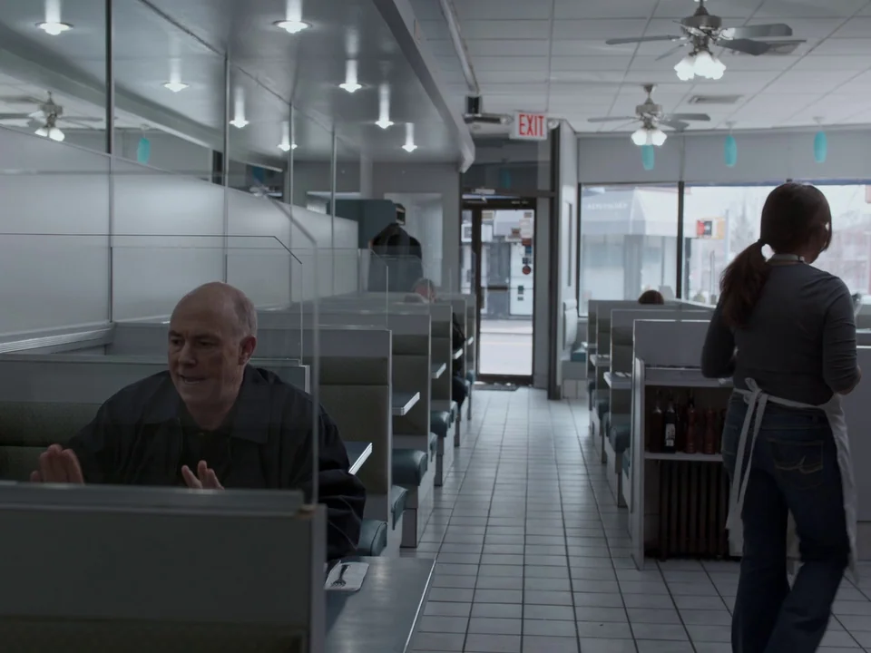 First Reformed (2017) movie still: medium — A diner booth with white acrylic partitions separates a seated man from the aisle. A waitress walks down…