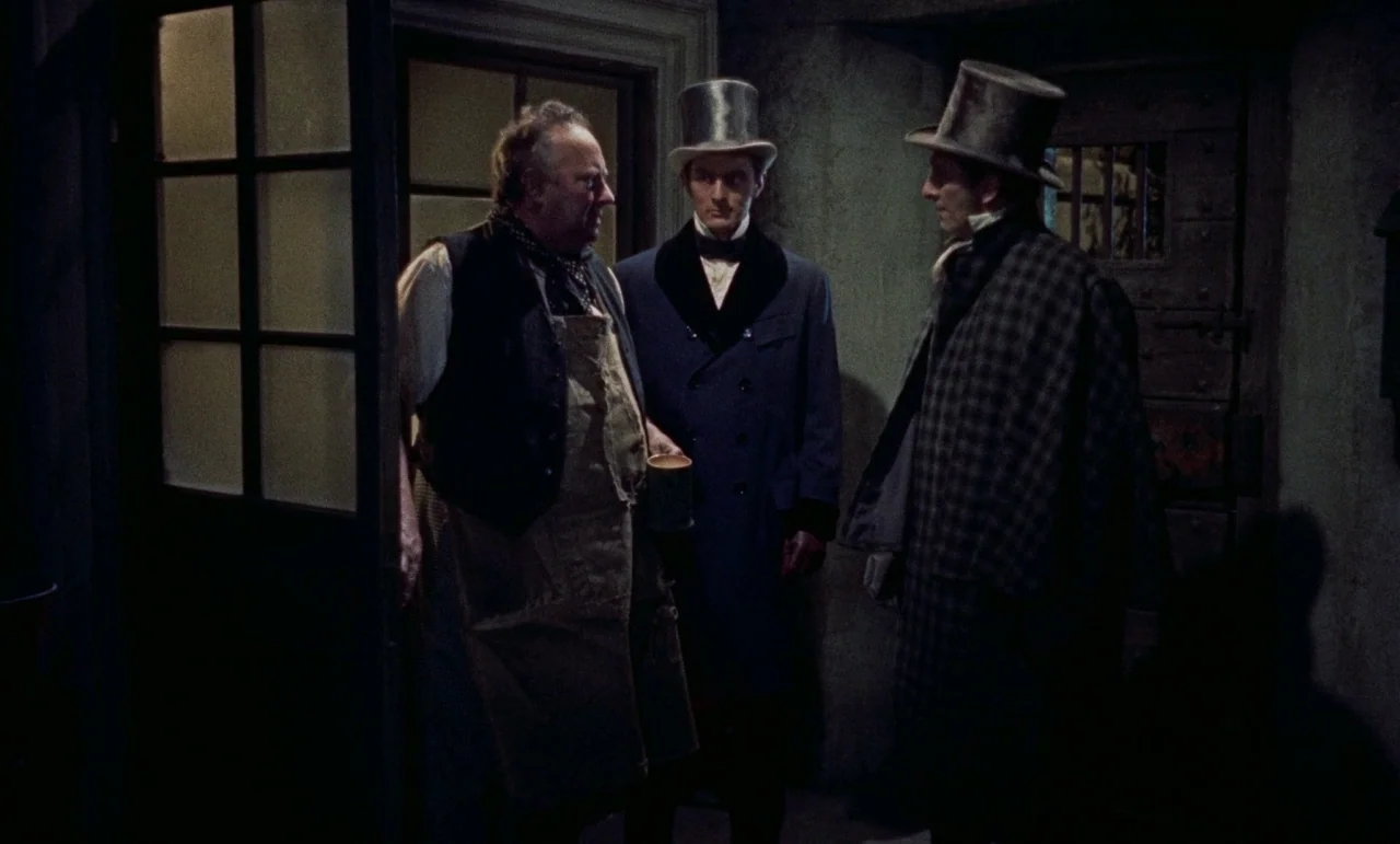 The Revenge of Frankenstein movie still: medium — A dimly lit room with stone walls and a wooden door featuring a small window. Three men in…
