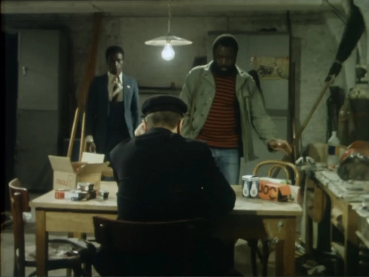 A Dessert for Constance (1981) movie still: medium — A cluttered workshop contains a wooden table with boxes and various industrial tools. Two men…