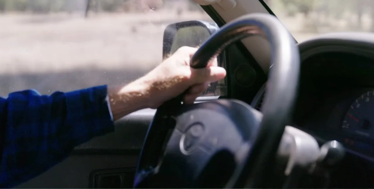 Alien Abduction: Travis Walton (2022) movie still: closeup — The interior of a moving vehicle shows the steering wheel and a driver's arm. A person…