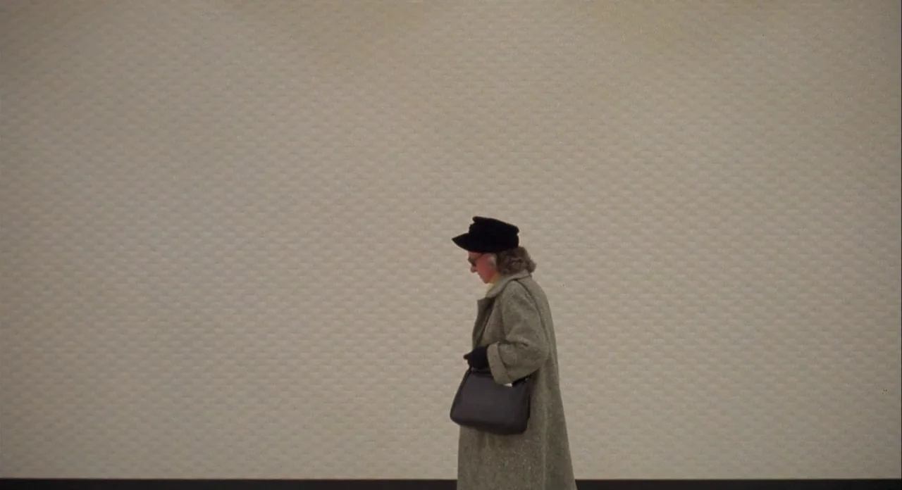 Koyaanasquatsi (1982) movie still: wide — A person walks against a plain, off-white textured wall background. The subject moves from left to right…