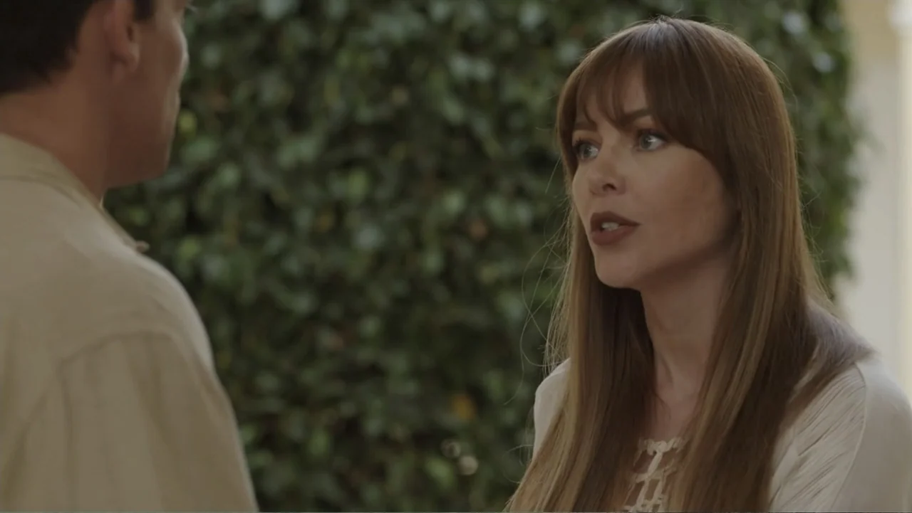 Por Amor al Money (2023) movie still: medium — An outdoor patio features dense green hedge foliage serving as a backdrop. A woman addresses a man…