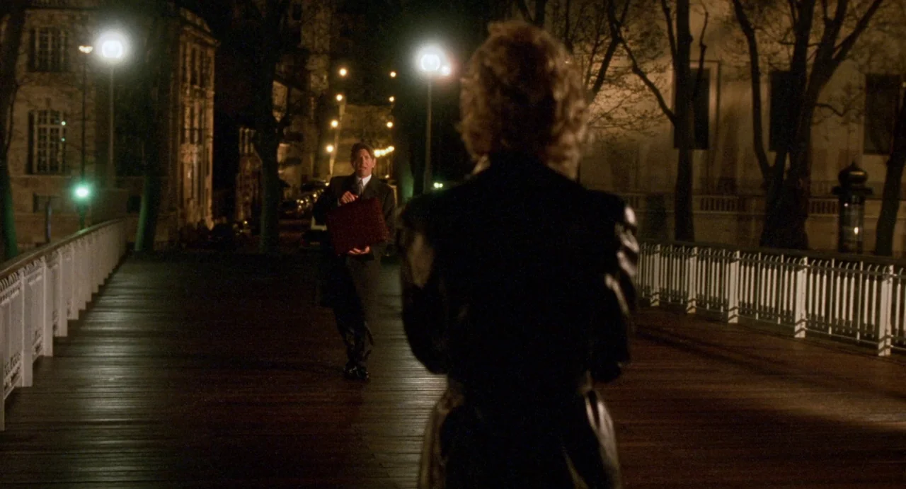 Femme Fatale (2002) movie still: medium — A wooden pedestrian bridge at night is illuminated by tall streetlights. A man with a briefcase approaches…