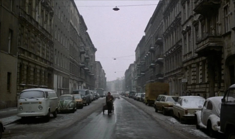 Stroszek (1977) movie still: wide — A dense urban street in winter is flanked by tall, darkened apartment blocks. A single person slowly pulls a…