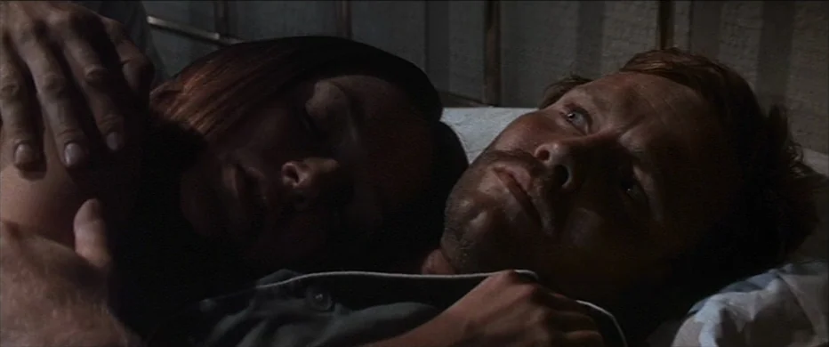 Deliverance (1972) movie still: closeup — A dark, dimly lit indoor space with two people lying in a bed. The man is awake while the woman sleeps…