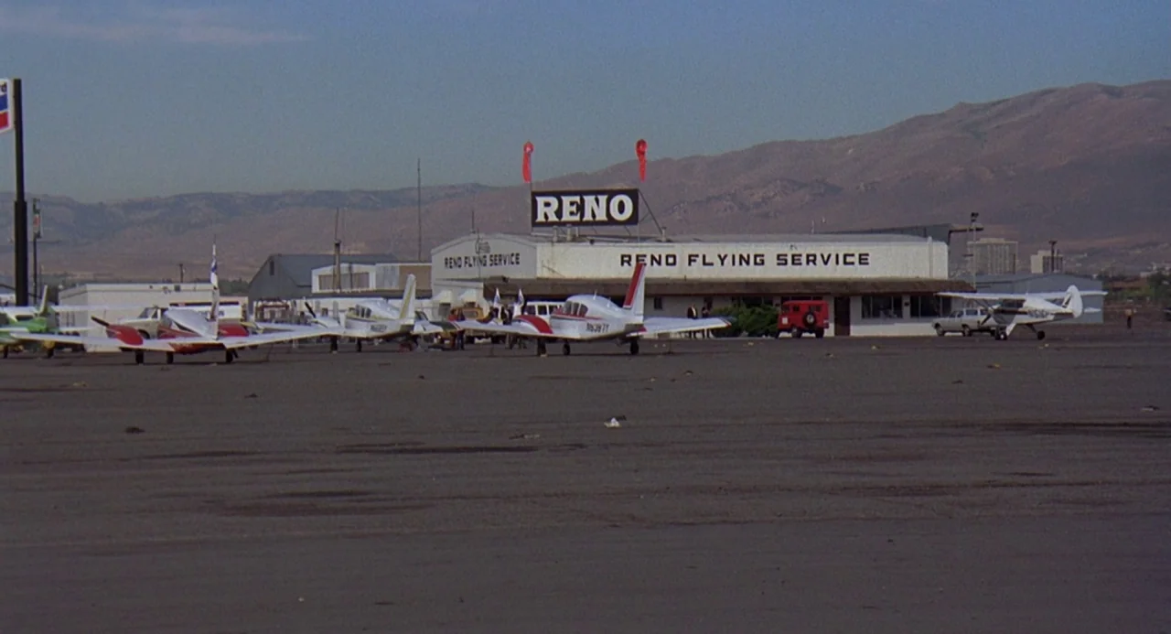 Charley Varrick (1973) movie still: wide — A flat, paved airfield sits before a low-slung building with Reno Flying Service signage. Mountains…