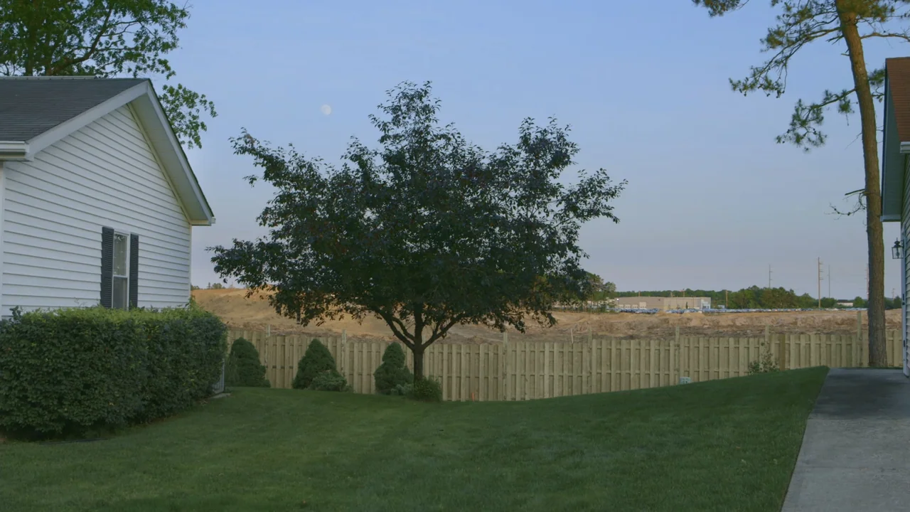 The Last Race (2022) movie still: wide — A residential backyard overlooks an adjacent plot of cleared earth and construction. A tree stands in the…