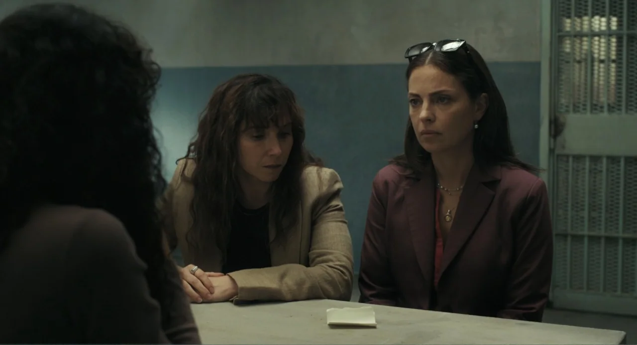 Belén (2025) movie still: medium — A austere room features pale blue walls and a metal barred door in back. Two women sit at a grey table looking…