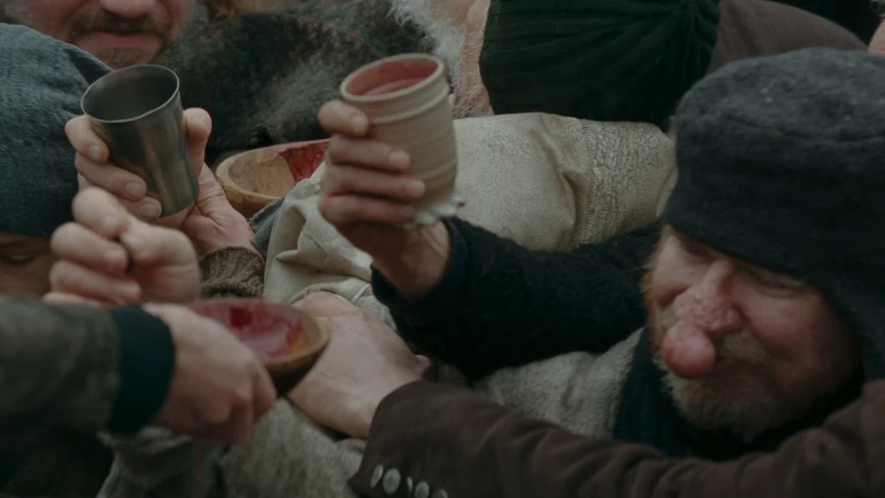 The Devil’s Bath (2024) movie still: medium — A group of people gathers together holding various drinking vessels in a cluttered environment. The…