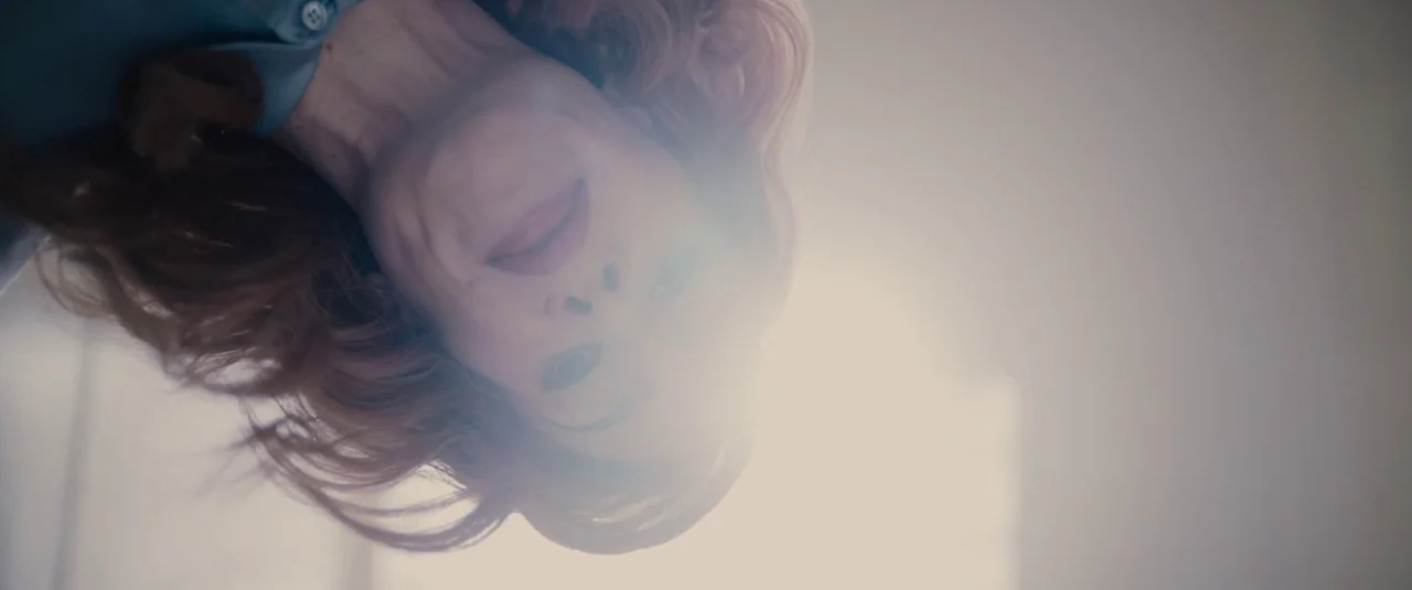 Glass (2019) movie still: closeup — A light-filled room with an off-screen light source creating high-key brightness. A woman with red hair looks…