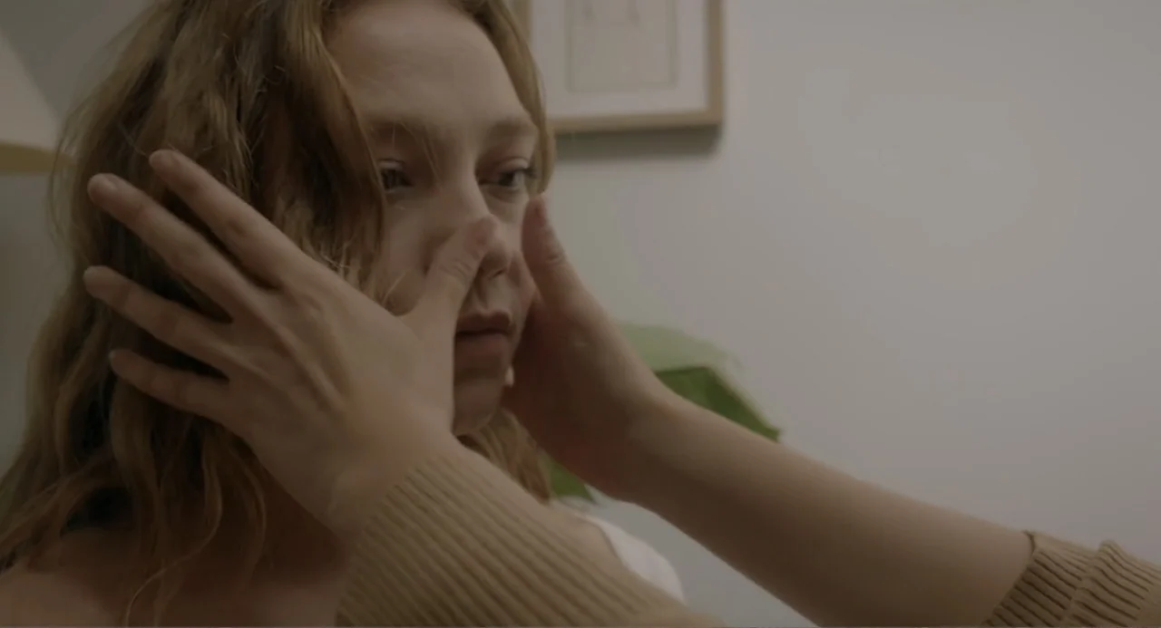 This Closeness (2024) movie still: closeup — A minimalist white room with a framed picture on the wall. A person's hands frame the face of a young…