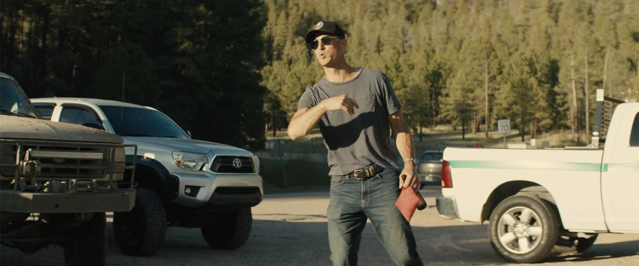 Those Who Wish Me Dead (2021) movie still: medium — A gravel parking area bordered by a pine-covered hillside during late afternoon light. A man in…